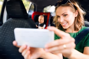 Young mother with her little son in the car. Woman taking selfie with a smartphone.