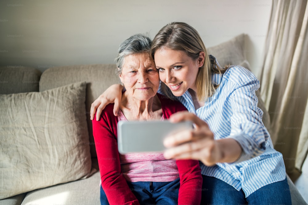 연로한 할머니와 성인 손녀가 집에서 스마트폰을 들고 소파에 앉아 셀카를 찍고 있다.