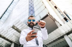 Une vue en contre-plongée d’un homme d’affaires hipster avec des lunettes de soleil et un smartphone debout dans la rue à Londres, prenant un selfie. Espace de copie.