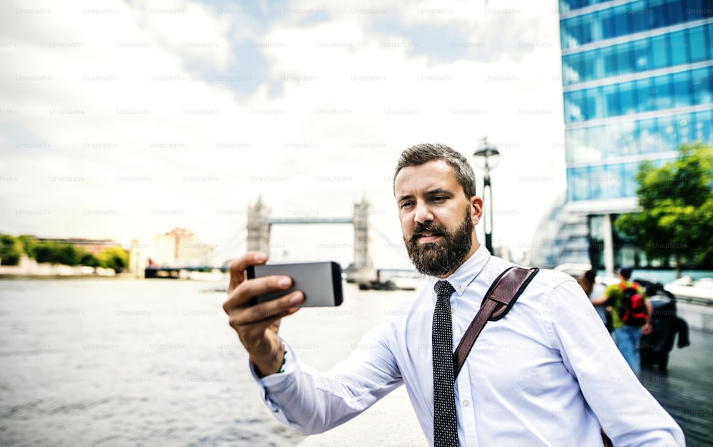 Hipster businessman with smartphone standing by the river Thames in London, taking selfie. Copy space.