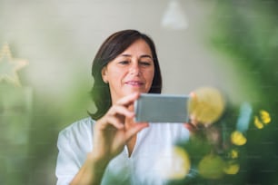 A senior woman with smartphone standing by a Christmas tree at home, taking selfie.