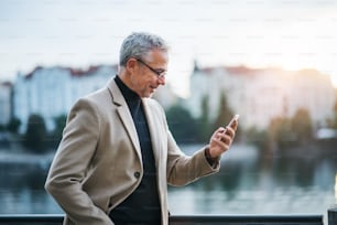 Mature handsome businessman with smartphone standing by river Vltava in Prague city at sunset, taking selfie. Copy space.