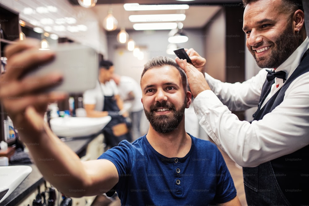 Cliente homem hipster bonito visitando haidresser e cabeleireiro em barbearia, tirando selfie com smartphone.