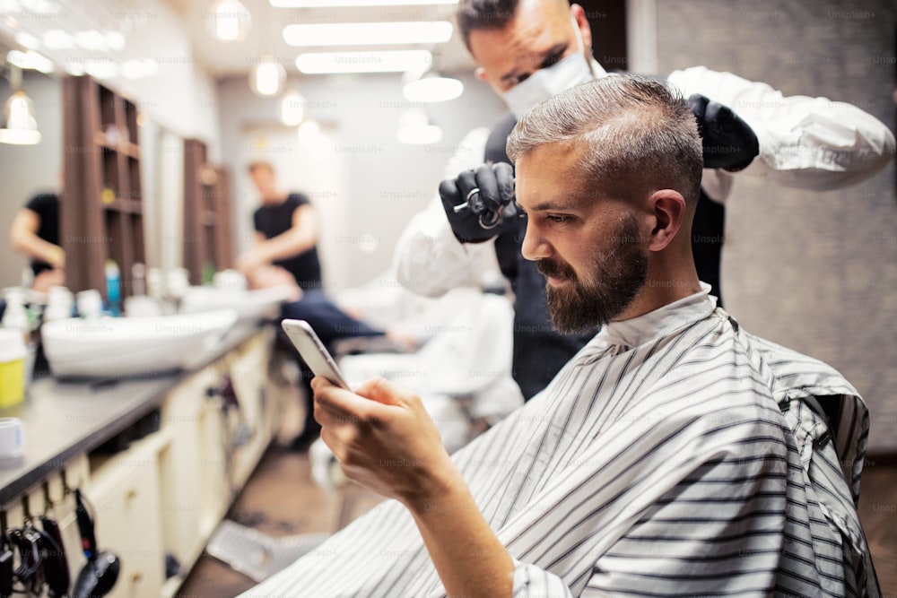 Guapo cliente hipster visitando a un gallinero y estilista en una peluquería, tomando selfies con un teléfono inteligente.