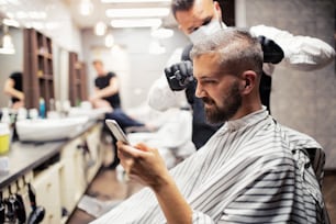 ハンサムなヒップスターの男性クライアントが理髪店のハイドレ�ッサーとヘアスタイリストを訪問し、スマートフォンで自分撮りをする。