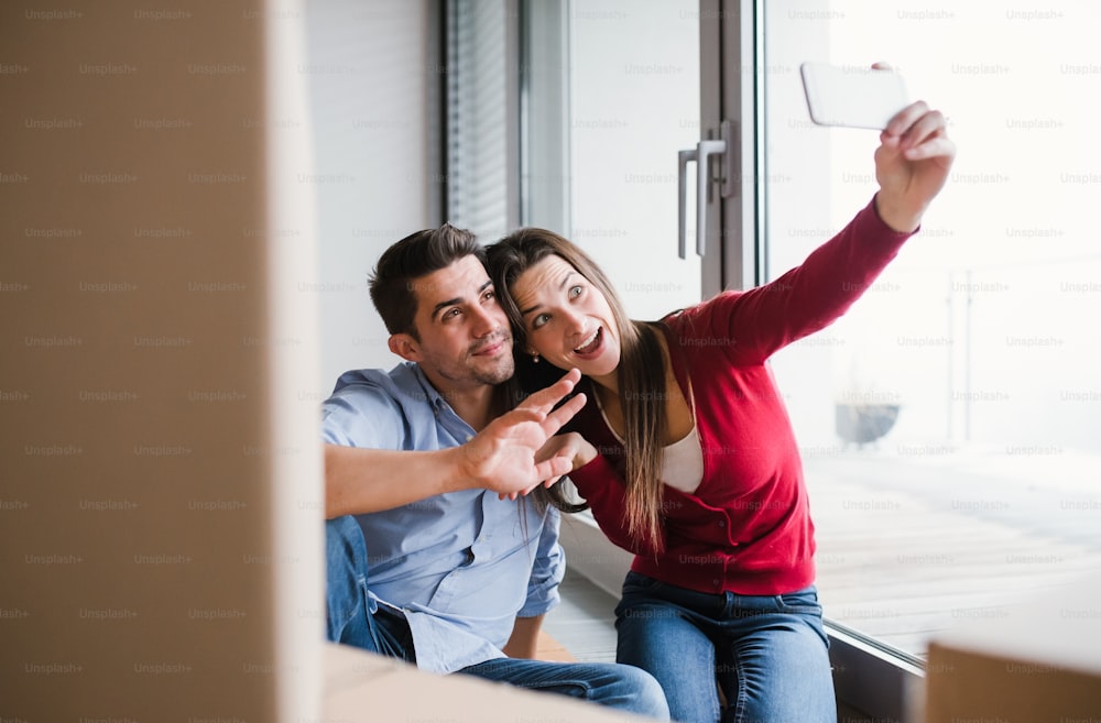 段ボール箱とスマートフォンを持つ若い夫婦が新しい家に引っ越し、自撮りをする。