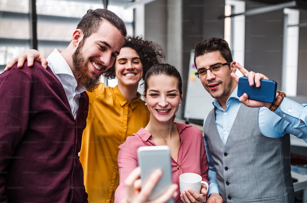Un grupo de jóvenes empresarios alegres con smartphone en la oficina, tomándose selfie.