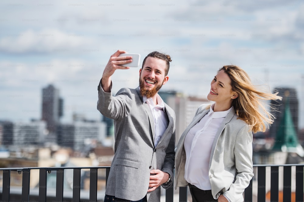 Dos alegres jóvenes empresarios con teléfono inteligente parados en una terraza fuera de la oficina, tomándose selfie.