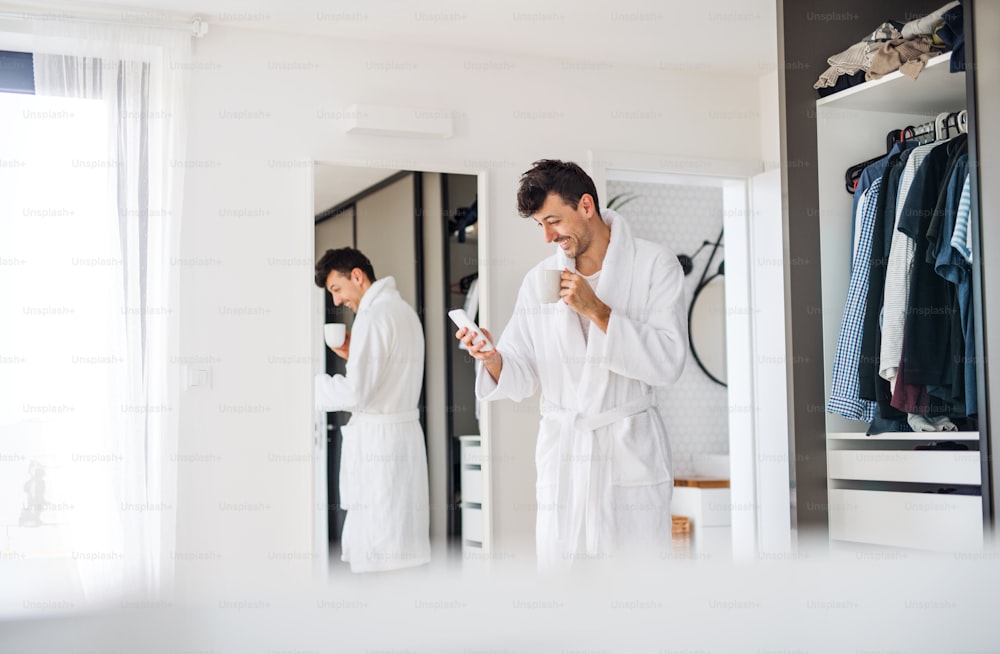 A young man with coffee and smartphone in the bedroom, a morning routine.