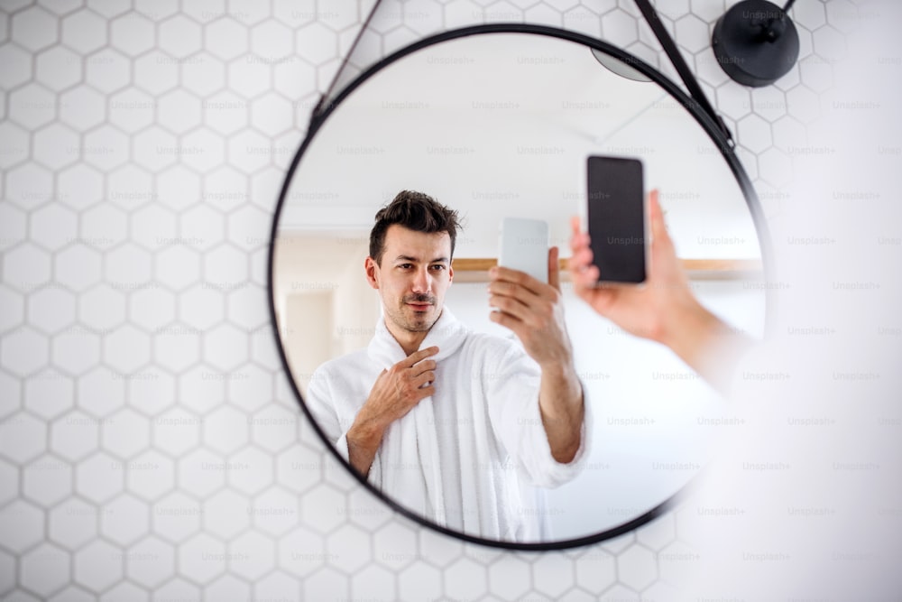 Young man with smartphone standing in the bathroom in the morning, taking selfie.