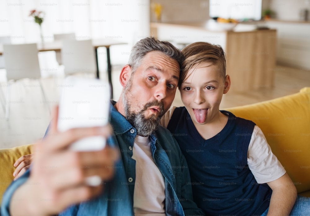 Père mûr avec un petit fils assis sur un canapé à l’intérieur, grimaçant en prenant un selfie.