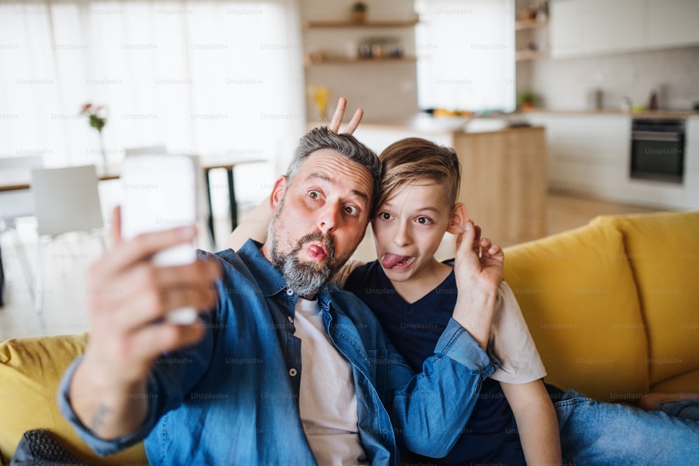 Père mûr avec un petit fils assis sur un canapé à l’intérieur, grimaçant en prenant un selfie.