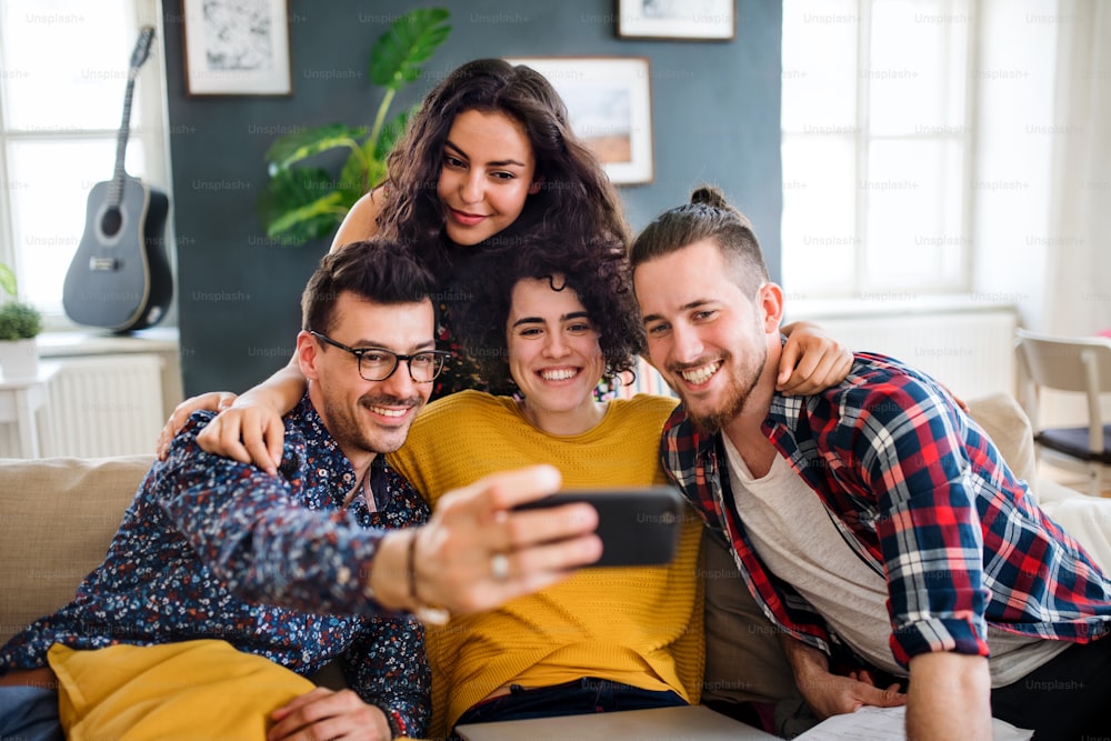 Un grupo de jóvenes amigos alegres con teléfono inteligente sentados en el sofá en el interior, tomando selfie. Concepto de casa compartida.