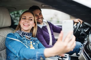 Una pareja joven feliz con un teléfono inteligente sentado en el automóvil, tomándose selfie.