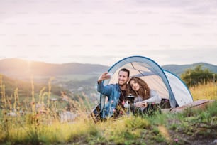 A young tourist couple travellers with tent shelter sitting in nature at sunset, taking selfie.