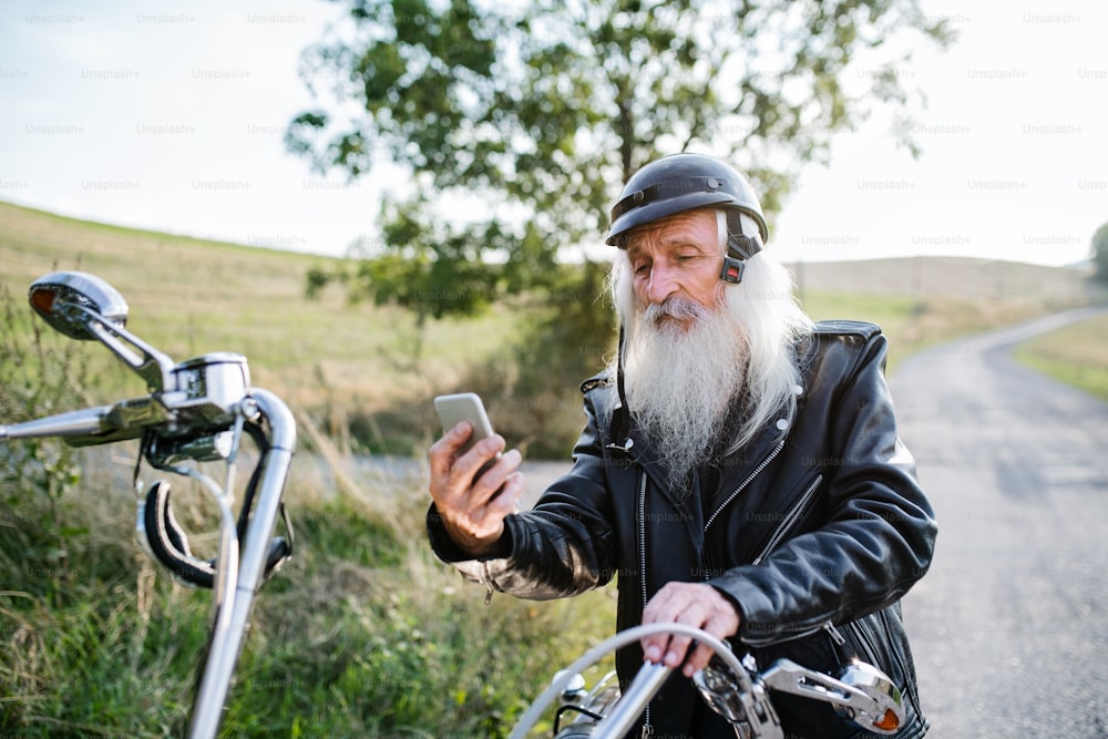 Un viajero mayor con una moto en el campo, tomándose selfie con un teléfono inteligente.