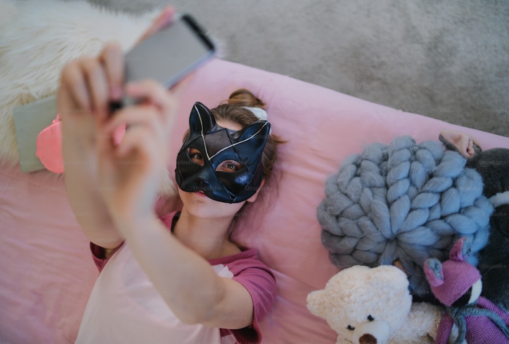 Top view of young girl with cat mask on bed, taking selfie. Online dating concept.