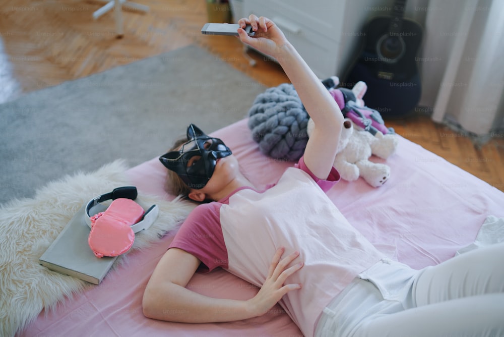 Top view of young girl with cat mask on bed, taking selfie. Online dating concept.