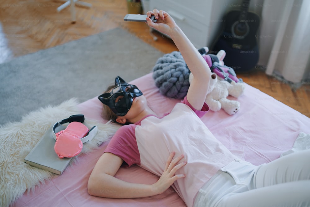 Vista superior de una niña con máscara de gato en la cama, tomando una selfie. Concepto de citas en línea.