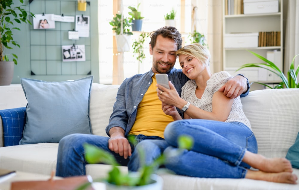 Vista frontal de una pareja feliz enamorada sentada en el interior de su casa, usando un teléfono inteligente.
