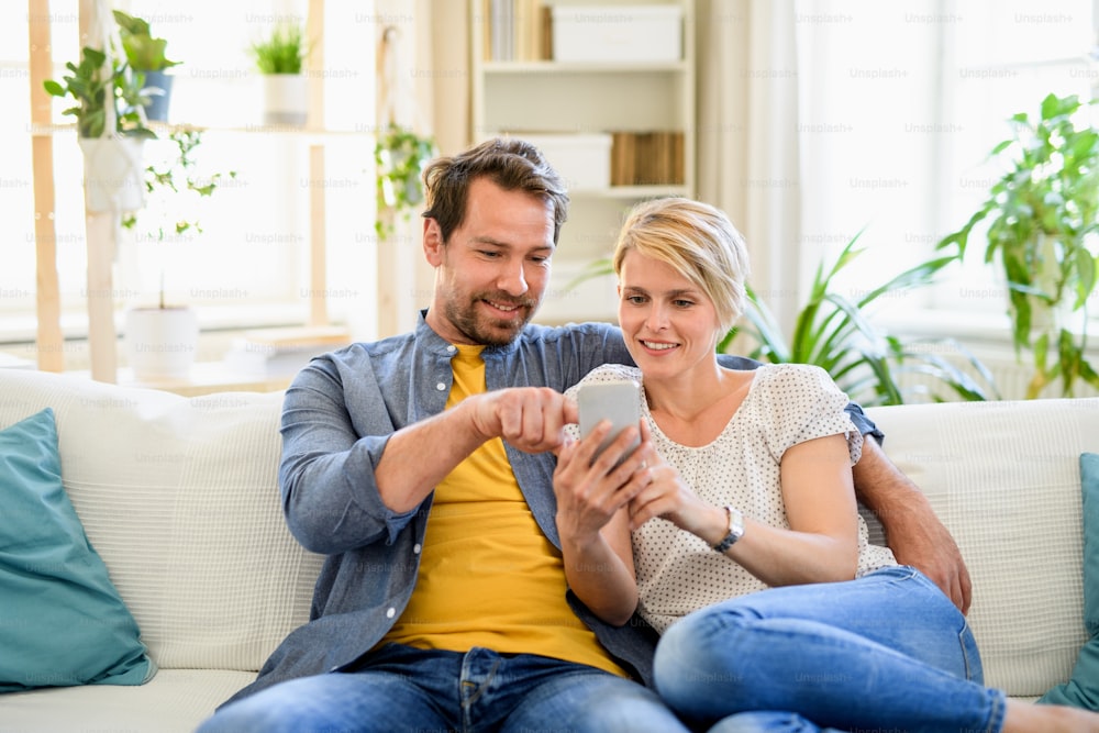 Vista frontal de una pareja feliz enamorada sentada en el interior de su casa, usando un teléfono inteligente.