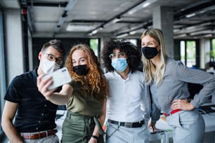 Young people with face masks back at work in office after coronavirus quarantine and lockdown, taking selfie.