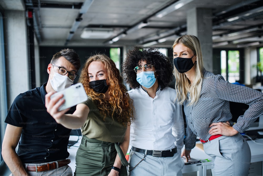 Young people with face masks back at work in office after coronavirus quarantine and lockdown, taking selfie.