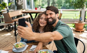 Casal feliz sentado ao ar livre no restaurante do terraço, tirando selfie com smartphone.