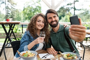 Happy couple sitting outdoors on terrace restaurant, taking selfie with smartphone.