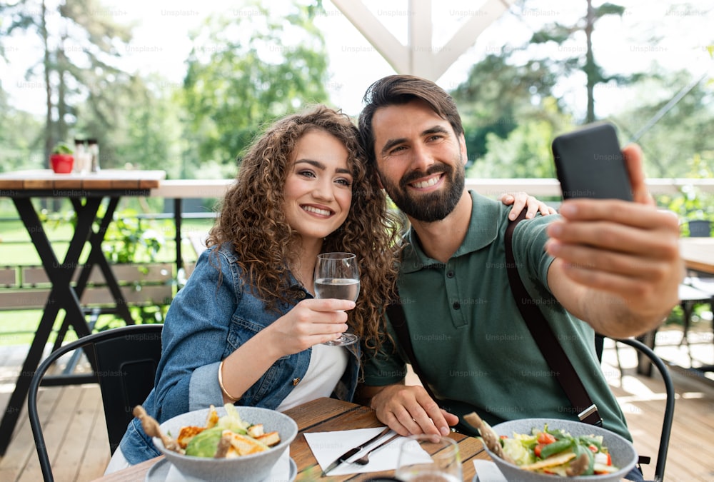 Coppia felice seduta all'aperto sul ristorante con terrazza, scattando selfie con lo smartphone.