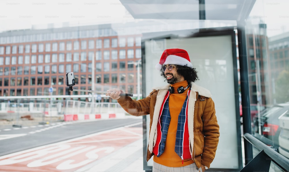 Young man with Santa hat making video for social media outdoors on bus stop. Copy space.