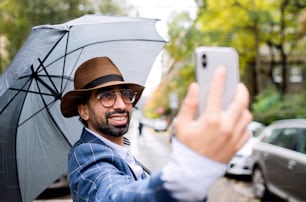 路上で屋外でソーシャルメディア用のビデオを作る傘を持つ若い男性のポートレート。
