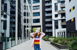 Front view portrait of young woman standing outdoors by block of flats in city.