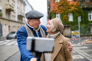 Portrait of happy senior couple walking outdoors on street in city, taking selfie.