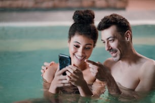 Portrait of happy young couple in indoor hot spring thermal pool, taking selfie.