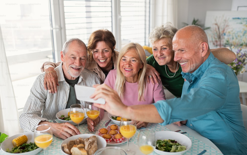 Um grupo de amigos idosos fazendo festa dentro de casa, tirando selfie ao comer na mesa.