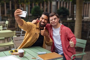 Un joven con síndrome de Down y su amigo mentor sentados y tomándose selfies al aire libre en un café