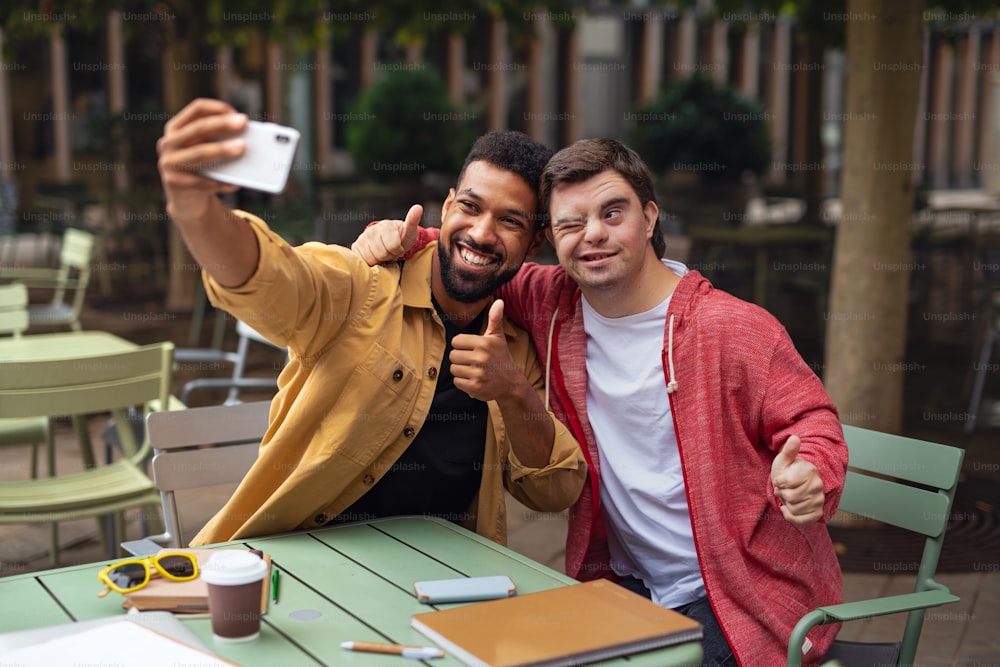 A young man with Down syndrome and his mentoring friend sitting and taking selfie outdoors in cafe
