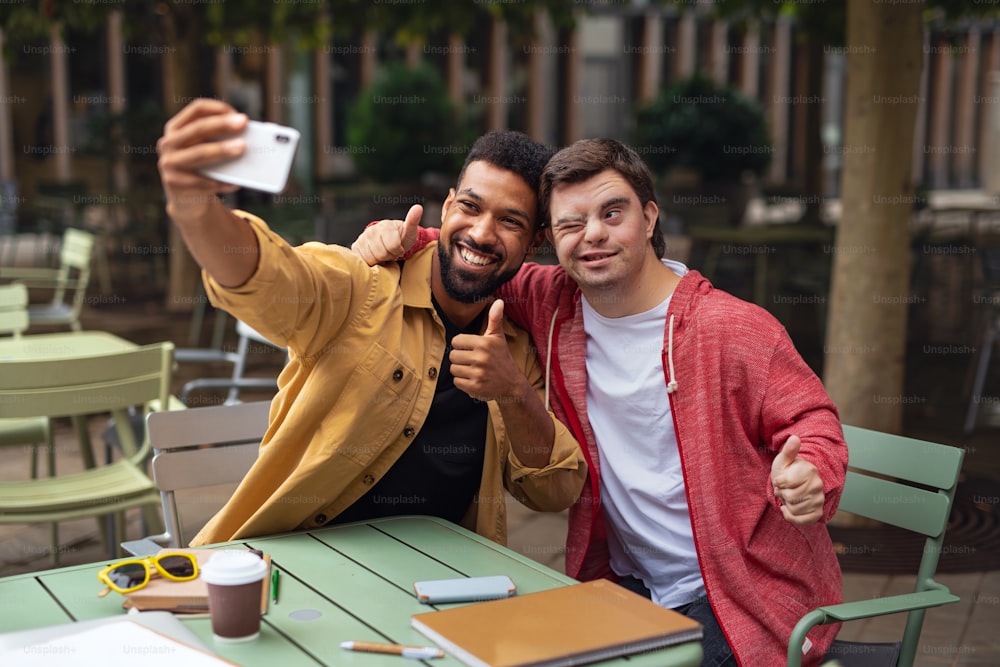 Ein junger Mann mit Down-Syndrom und sein Mentoring-Freund sitzen und machen Selfies im Freien im Café