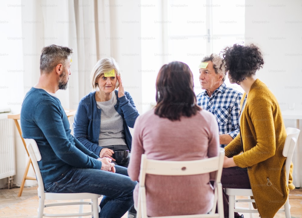 Men and women sitting in a circle during group therapy, adhesive notes with negative emotions on forehead.