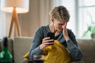 Worried woman with smartphone indoors on sofa at home feeling stressed, mental health concept.