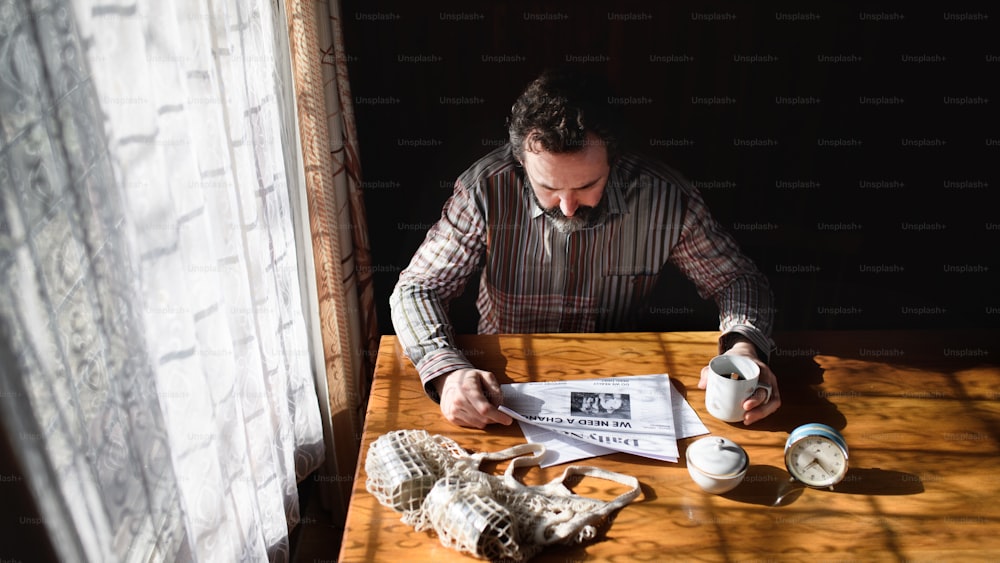 Portrait of unhappy poor mature man with coffee reading newspapers indoors at home, poverty concept.