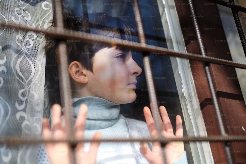 Povera bambina triste in piedi vicino alla finestra in casa a casa, concetto di povertà e coronavirus.