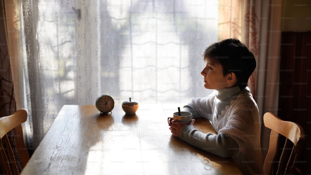 Side view of poor sad small girl with tea sitting at the table indoors at home, poverty concept.