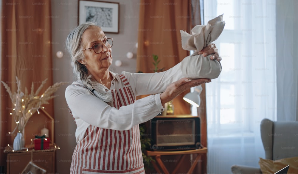 A senior woman holding Christmas present wrapped in eco materials indoors at home.