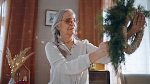 Mujer mayor una haciendo corona de Navidad con materiales naturales en el interior de casa