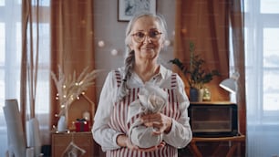 Eine ältere Frau, die zu Hause ein in Öko-Materialien gehülltes Weihnachtsgeschenk hält und in die Kamera schaut