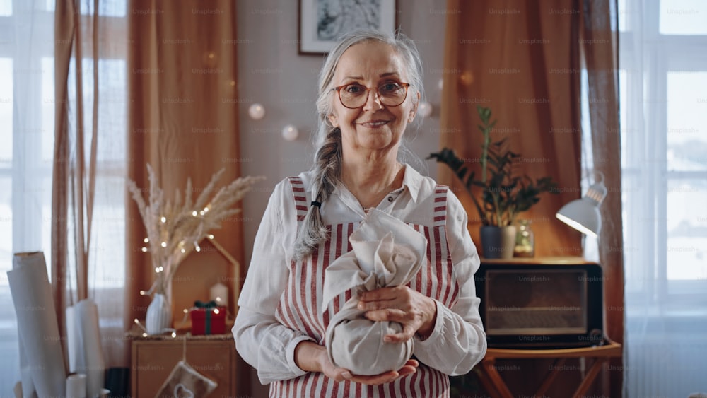 A senior woman holding Christmas present wrapped in eco materials indoors at home, looking at camera
