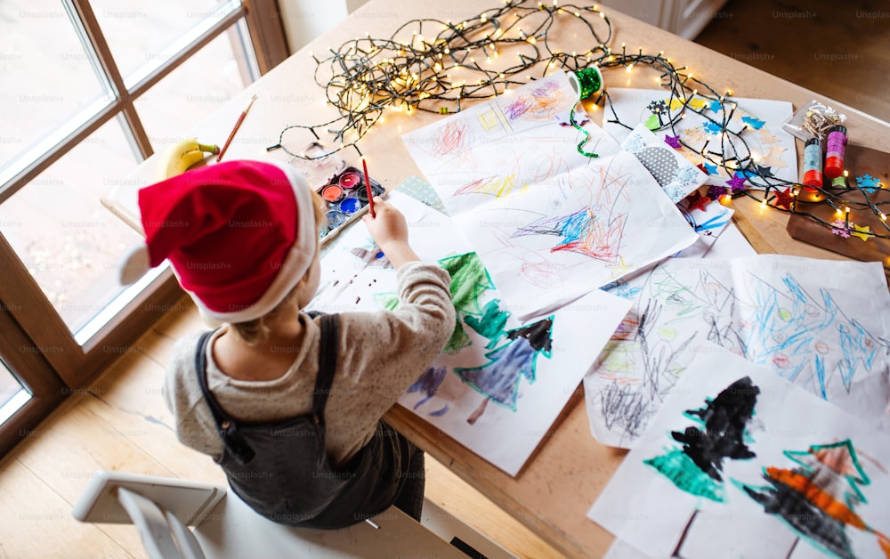Draufsicht auf ein nicht wiederzuerkennendes kleines Kind zu Hause zu Weihnachten, das Bilder mit Wasserfarben malt.