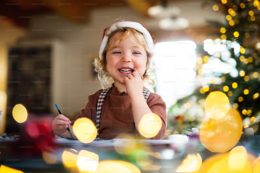 Portrait d’une petite fille heureuse à l’intérieur à la maison à Noël, peignant des tableaux.